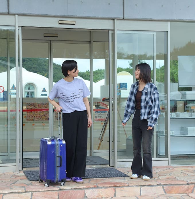 ガラス扉のエントランス前で会話する2人、左の人はスーツケースを持ち、右の人はカジュアルなシャツを着ている