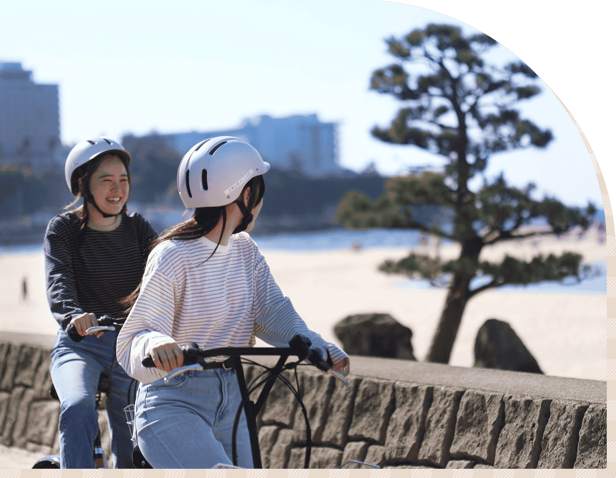ヘルメットを着用した2人が、海沿いの遊歩道で自転車に乗り、笑顔で会話しながら走っている様子
